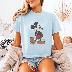 May include: Light blue t-shirt featuring a classic Mickey Mouse graphic. The cartoon character is depicted in black, red, and brown. The shirt is a short-sleeved, crew neck style.
