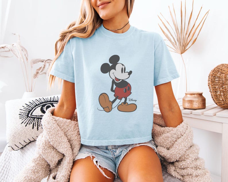 May include: Light blue t-shirt featuring a classic Mickey Mouse graphic. The cartoon character is depicted in black, red, and brown. The shirt is a short-sleeved, crew neck style.