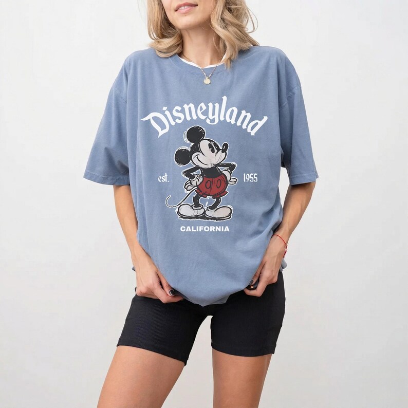 May include: A blue t-shirt with the Disneyland logo and a Mickey Mouse graphic. The graphic includes the text "est. 1955" and "CALIFORNIA". The shirt is a light blue color and has short sleeves.