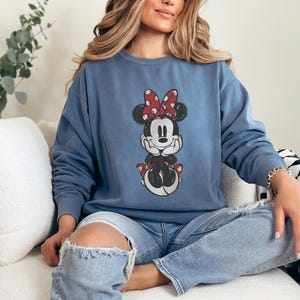 Puede incluir: Una sudadera azul de cuello redondo con un gr&aacute;fico de Minnie Mouse. El gr&aacute;fico muestra a Minnie Mouse con un lazo rojo con lunares blancos. La sudadera es de color azul liso y tiene mangas largas.