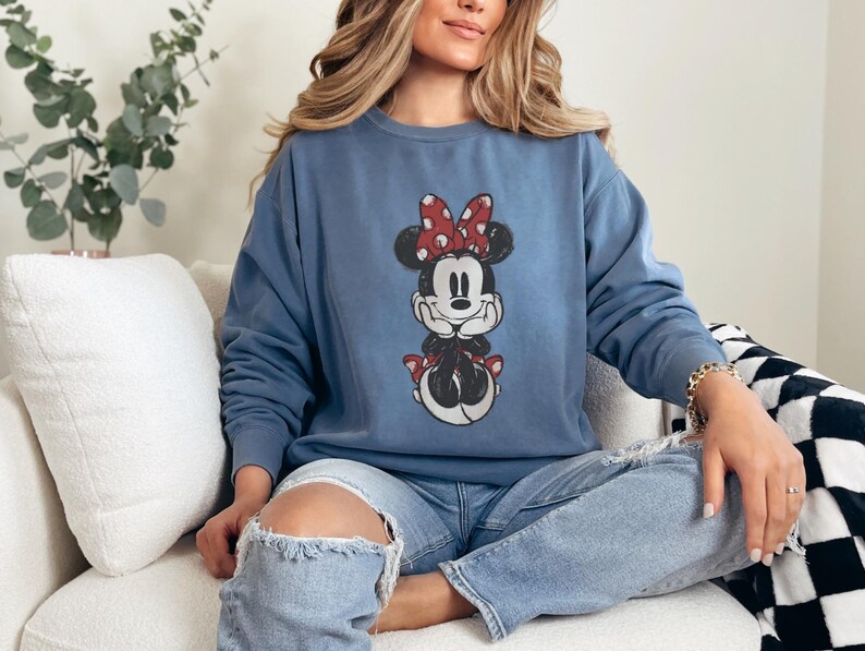 Puede incluir: Una sudadera azul de cuello redondo con un gr&aacute;fico de Minnie Mouse. El gr&aacute;fico muestra a Minnie Mouse con un lazo rojo con lunares blancos. La sudadera es de color azul liso y tiene mangas largas.