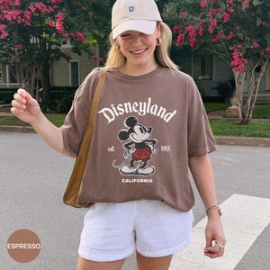 May include: Espresso-colored t-shirt with a Disneyland graphic featuring Mickey Mouse. The shirt has the text "Disneyland est. 1955 CALIFORNIA" in white. The person is wearing white shorts and a beige baseball cap.