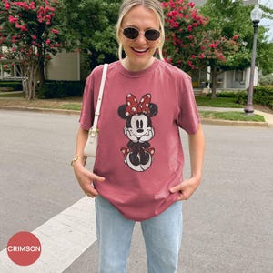 Puede incluir: Camiseta de color carmes&iacute; con un gr&aacute;fico de Minnie Mouse. El gr&aacute;fico muestra a Minnie Mouse con un lazo rojo con lunares blancos. La camiseta se combina con vaqueros azul claro. La palabra "CRIMSON" est&aacute; en un c&iacute;rculo rojo en la esquina inferior izquierda.