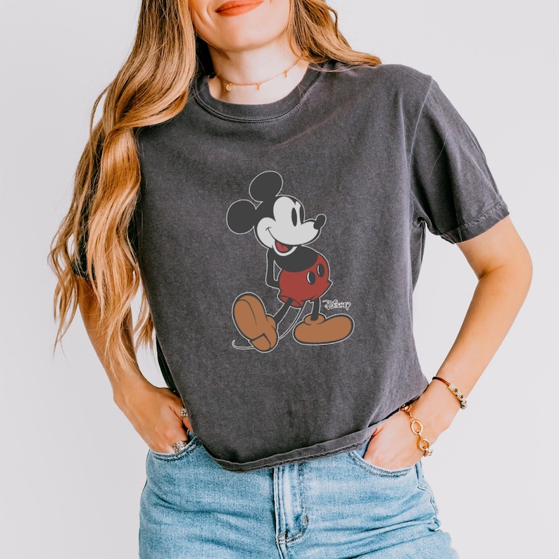 May include: Dark gray t-shirt featuring a classic Mickey Mouse graphic. The cartoon character is depicted in black, red, and brown, with the Disney logo. The shirt has a vintage, worn look.