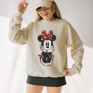May include: Beige sweatshirt featuring a Minnie Mouse graphic. The graphic shows Minnie Mouse with a red polka dot bow, black ears, and a red skirt. The sweatshirt has long sleeves and a crew neck. The person in the image is wearing a beige cap and sunglasses.
