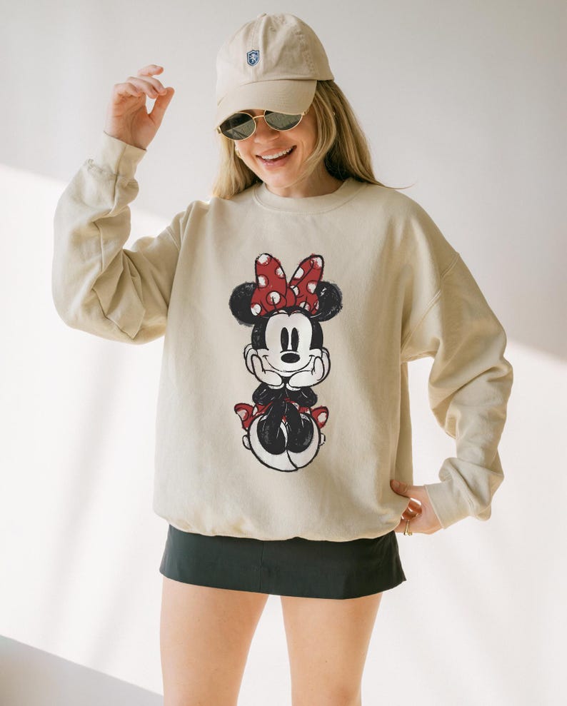 May include: Beige sweatshirt featuring a Minnie Mouse graphic. The graphic shows Minnie Mouse with a red polka dot bow, black ears, and a red skirt. The sweatshirt has long sleeves and a crew neck. The person in the image is wearing a beige cap and sunglasses.
