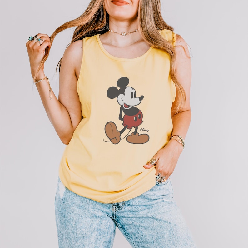 May include: Yellow tank top featuring a classic Mickey Mouse graphic. The cartoon character is depicted in black, red, and brown. The tank top is a solid yellow color and has a round neckline. The Disney logo is visible.