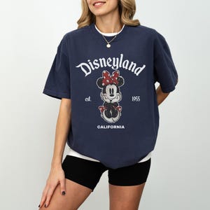 May include: Navy blue t-shirt with a white collar featuring the Disneyland logo and a Minnie Mouse graphic. The graphic includes the text "est. 1955" and "CALIFORNIA". The shirt is a relaxed fit.