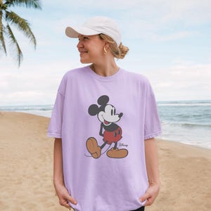 May include: A light purple short-sleeved t-shirt featuring a graphic print of Mickey Mouse in his classic black, white, and red color scheme, with the word Disney printed in black script next to the character's feet and a white baseball cap.
