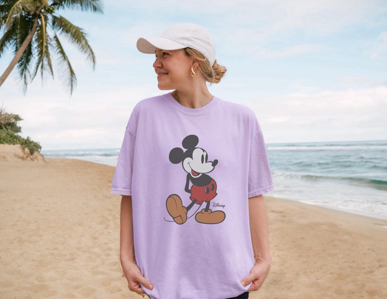 May include: A light purple short-sleeved t-shirt featuring a graphic print of Mickey Mouse in his classic black, white, and red color scheme, with the word Disney printed in black script next to the character's feet and a white baseball cap.