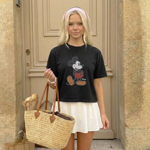 May include: Black Mickey Mouse graphic t-shirt paired with a white skirt. A straw tote bag with brown leather handles and a pink headband complete the look. The t-shirt features a classic Mickey Mouse design.