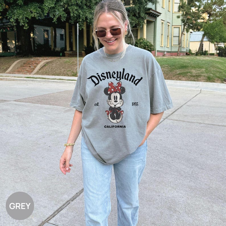 May include: A grey t-shirt with the Disneyland logo and a Minnie Mouse graphic. The text "Disneyland est. 1955 CALIFORNIA" is printed on the front. The shirt is paired with light blue jeans.