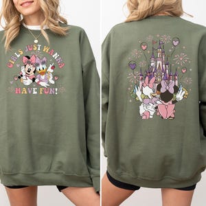May include: An olive green sweatshirt with a Minnie Mouse and Daisy Duck design and the text "GIRLS JUST WANNA HAVE FUN!". The back features a castle, fireworks, and the two characters. The design is in pink, purple, and white.
