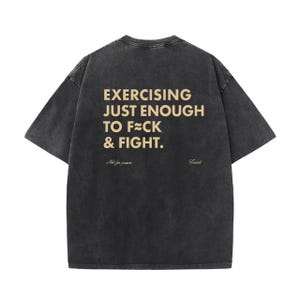 Può includere: Una maglietta grigio scuro oversize con il testo dorato "EXERCISING JUST ENOUGH TO F**K & FIGHT". La maglietta presenta anche le frasi "Not for peace" e "Found".