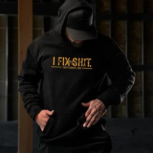 May include: Black hooded sweatshirt with the text "I FIX SHIT." in a distressed gold font. Below the text is the phrase "THAT'S WHAT I DO" in a smaller gold font. The sweatshirt has a front pocket and a black cap.
