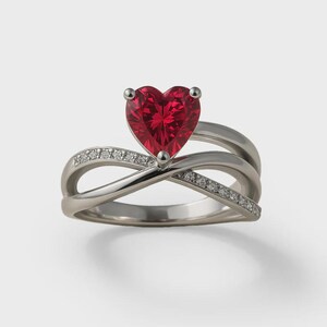 Op de afbeelding: Een zilverkleurige ring met een opvallende hartvormige rode edelsteen in het midden. De ringband bestaat uit twee in elkaar verstrengelde metalen strengen, waarvan één streng is bezet met een rij kleine, heldere, fonkelende steentjes.
