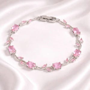Op de afbeelding: Een zilveren armband met ovale roze edelstenen en kleine roze bladvormige details. De armband heeft een delicaat, bloemig ontwerp en een veilige sluiting. De edelstenen zijn gezet in zilver, wat een vrouwelijk en elegant accessoire creëert.