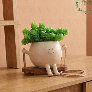 Puede incluir: Una maceta de cerámica blanquecina con forma de personaje sonriente, sosteniendo un columpio de madera. La maceta está llena de plantas suculentas verdes. El columpio está suspendido por una cuerda. El fondo es de color melocotón claro.