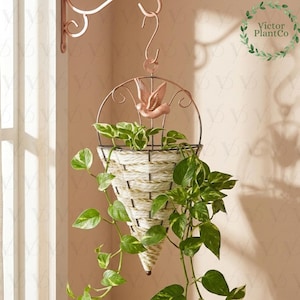 May include: A hanging planter with a woven basket design, filled with green plants. The planter is suspended from a decorative metal bracket with a bird accent. The basket is white and tan, and the metal is a copper colour.