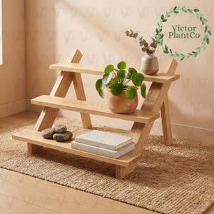 Pode incluir: Um suporte para plantas de madeira clara de três níveis com design em forma de A. O suporte contém uma planta Pilea peperomioides num vaso de terracota, um pequeno vaso de cerâmica com vegetação, dois livros empilhados e três pedras cinzentas.