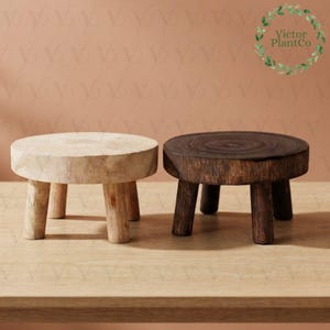 May include: Two round wooden plant stands with three legs each. One stand is light natural wood, and the other is dark stained wood. Both feature a visible circular grain pattern on the top surface. The logo Victor PlantCo is in the corner.