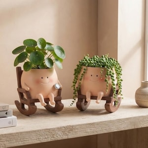 May include: Two light-skinned, round-shaped planters with smiling faces, each sitting in a small brown wooden rocking chair. One planter holds a Pilea peperomioides plant, while the other contains a trailing string of pearls succulent plant.
