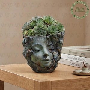 May include: A sculptural planter shaped like a human face with closed eyes, finished in a weathered blue-green patina. The vessel is filled with a cluster of green succulent plants. The text Victor Plant Co is visible in the upper right corner.
