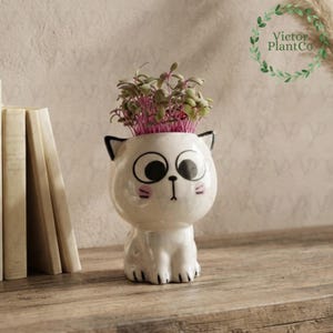 May include: A white ceramic cat-shaped planter with large black eyes, a small nose, and pink cheek details. The planter holds a cluster of small, leafy green and purple sprouts. The cat figure has pointed ears and a rounded body shape.