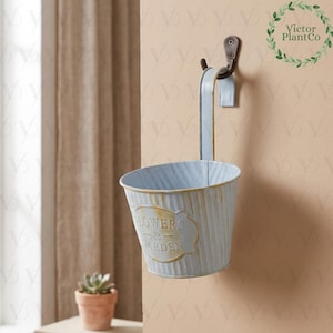 May include: A light blue metal hanging planter with a distressed finish, featuring the text "Flowers & Garden". The planter is suspended from a dark metal hook, ideal for displaying small plants or flowers. The planter is approximately 15 cm in diameter.