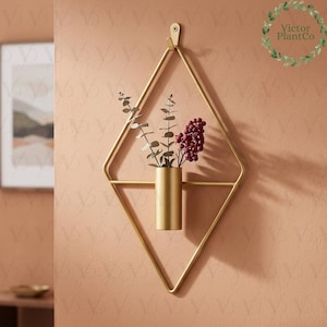 Puede incluir: Un macetero de metal dorado montado en la pared con forma de diamante geométrico. El macetero contiene un pequeño arreglo de vegetación y bayas de color burdeos. El macetero está colgado en una pared de color melocotón.