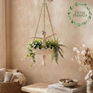 May include: A hanging planter featuring a cream-colored figurine of a dancer with arms outstretched, positioned in the center of a scalloped bowl. The planter is suspended by three gold-toned metal chains and contains green leafy plants.