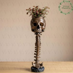 May include: A bronze-colored skull-shaped planter with a spine-like stem and a black base. The planter holds a small plant with red and green leaves. The planter has the text "Victor PlantCo" in a circular design.