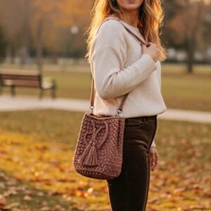 May include: A crocheted shoulder bag in a dusty rose color with a tassel detail. The bag is worn over the shoulder. The person is wearing a cream-colored sweater and brown pants. The background is a park with autumn leaves.