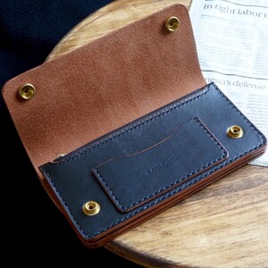 May include: A bifold wallet crafted from dark navy and tan leather, featuring four brass snap fasteners and a stitched card slot on the front. The interior displays a natural tan suede texture, while the exterior pocket is embossed with the text Millibie.