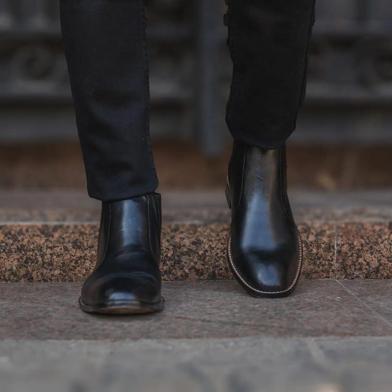 May include: A pair of black leather Chelsea boots. The boots have a polished finish and a classic design, with elastic side panels. The boots are worn with black pants, creating a sleek, monochromatic look.