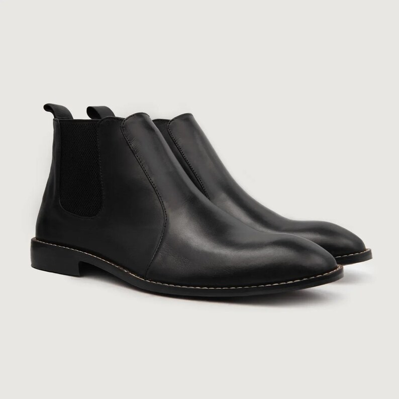 May include: A pair of black leather Chelsea boots. The boots have a rounded toe, elastic side panels, and a pull tab at the back. The soles are stitched with a light-colored thread. The boots are displayed on a white background.