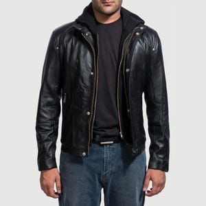 May include: A black leather jacket with a zip-up front and a hooded sweatshirt underneath. The jacket has a classic biker style with a notched collar and multiple pockets. The model is wearing blue jeans.