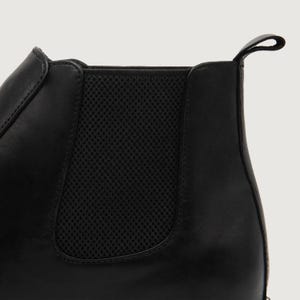 May include: Close-up of a black leather Chelsea boot. The boot features a pull tab at the back and elastic side panels for easy wear. The leather has a smooth finish, and the design is minimalist.