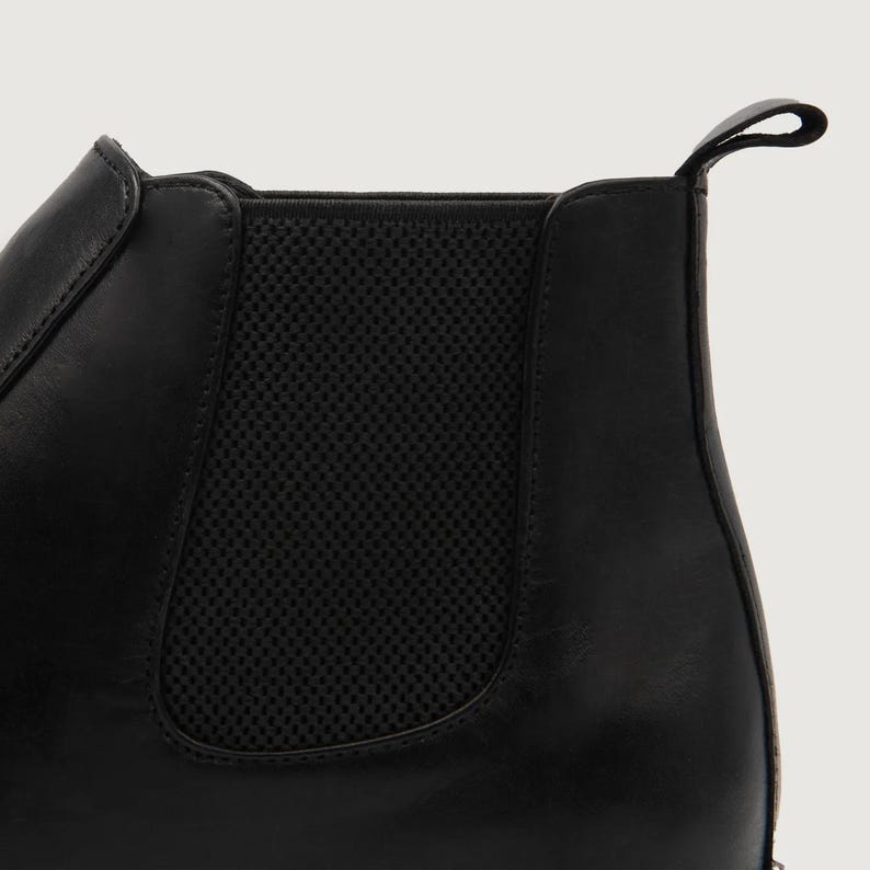 May include: Close-up of a black leather Chelsea boot. The boot features a pull tab at the back and elastic side panels for easy wear. The leather has a smooth finish, and the design is minimalist.