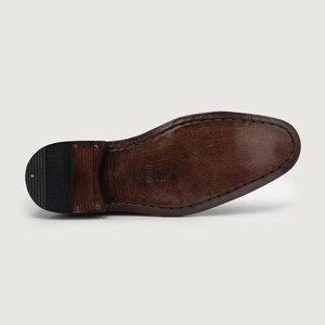 May include: Close-up of the sole of a brown leather shoe. The sole is a dark brown color with a textured surface and visible stitching. The heel is a darker shade of brown with a rubber tread.