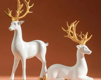 May include: Two white deer figurines with gold antlers and hooves. One deer stands, the other reclines. The figurines have a glossy finish and are set against a warm brown background. These decorative sculptures add a touch of elegance.