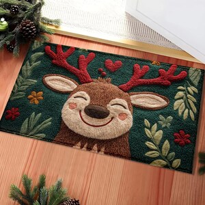 May include: A rectangular green doormat featuring a smiling reindeer with red antlers and a brown face. The mat is decorated with floral and leaf accents, and two red hearts. The doormat is placed on a wooden floor.