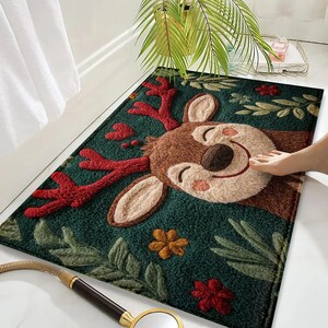 May include: A rectangular bath mat featuring a smiling reindeer design. The mat has a dark green background with red antlers, brown fur, and floral accents. The mat is placed on a white tiled floor.