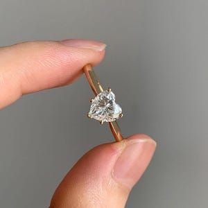 May include: A gold ring featuring a heart-shaped diamond. The diamond is clear and faceted, catching the light. The ring's band is a thin, polished gold. The ring is held between fingers against a neutral background.
