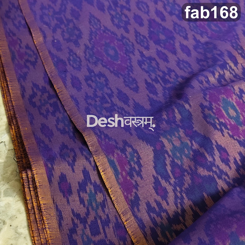 May include: A purple fabric featuring a repeating ikat pattern in darker shades of violet and blue. The textile has a visible sheen and a finished edge. The text fab168 and Deshवस्त्रम् are printed on the surface of the folded material.