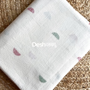 May include: White fabric featuring a pattern of half-circle shapes in red, green, and purple. The fabric has the text "Desh" and "fab124" printed on it. The fabric is folded and placed on a woven surface.