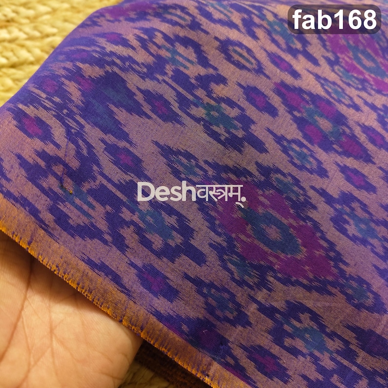 May include: A purple fabric featuring a repeating ikat-style pattern in shades of blue and magenta. The textile displays the text "fab168" in the top right corner and "Deshवस्त्रम्." in the center. The edge of the fabric has a thin orange border.