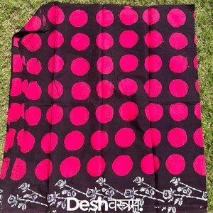 Puede incluir: Un textil negro con un patrón repetitivo de grandes círculos de color rosa brillante. La tela tiene un borde decorativo con diseños florales blancos y el texto "Desh वस्त्रम". La etiqueta "Fab079" es visible. El textil se muestra sobre una superficie verde.