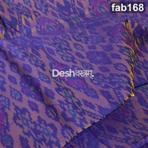 May include: A purple fabric featuring a repeating ikat pattern in shades of blue and violet. The textile displays the text fab168 in the top right corner and the word Deshवस्त्रम् in the center, printed in a clear white font on the surface.