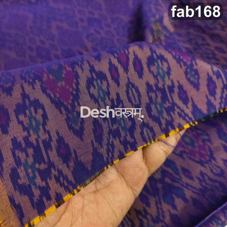May include: A purple fabric featuring a repeating ikat pattern in darker shades of blue and violet. The textile displays the text Deshवस्त्रम् and fab168. The edge of the fabric shows a thin yellow and black striped border detail.